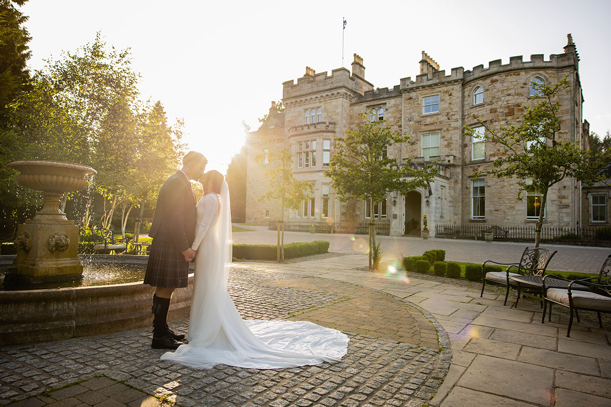 Bride and groom share a kiss beside a stone fountain at sunset, the castle glowing warmly behind them