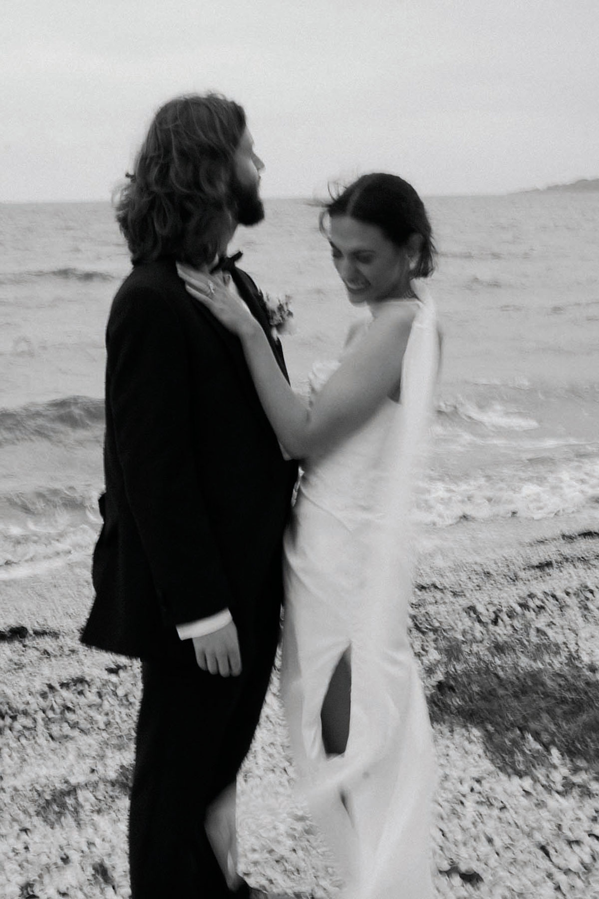 Bride and groom sharing a relaxed moment on a pebbled beach, with the sea behind them, captured in black and white