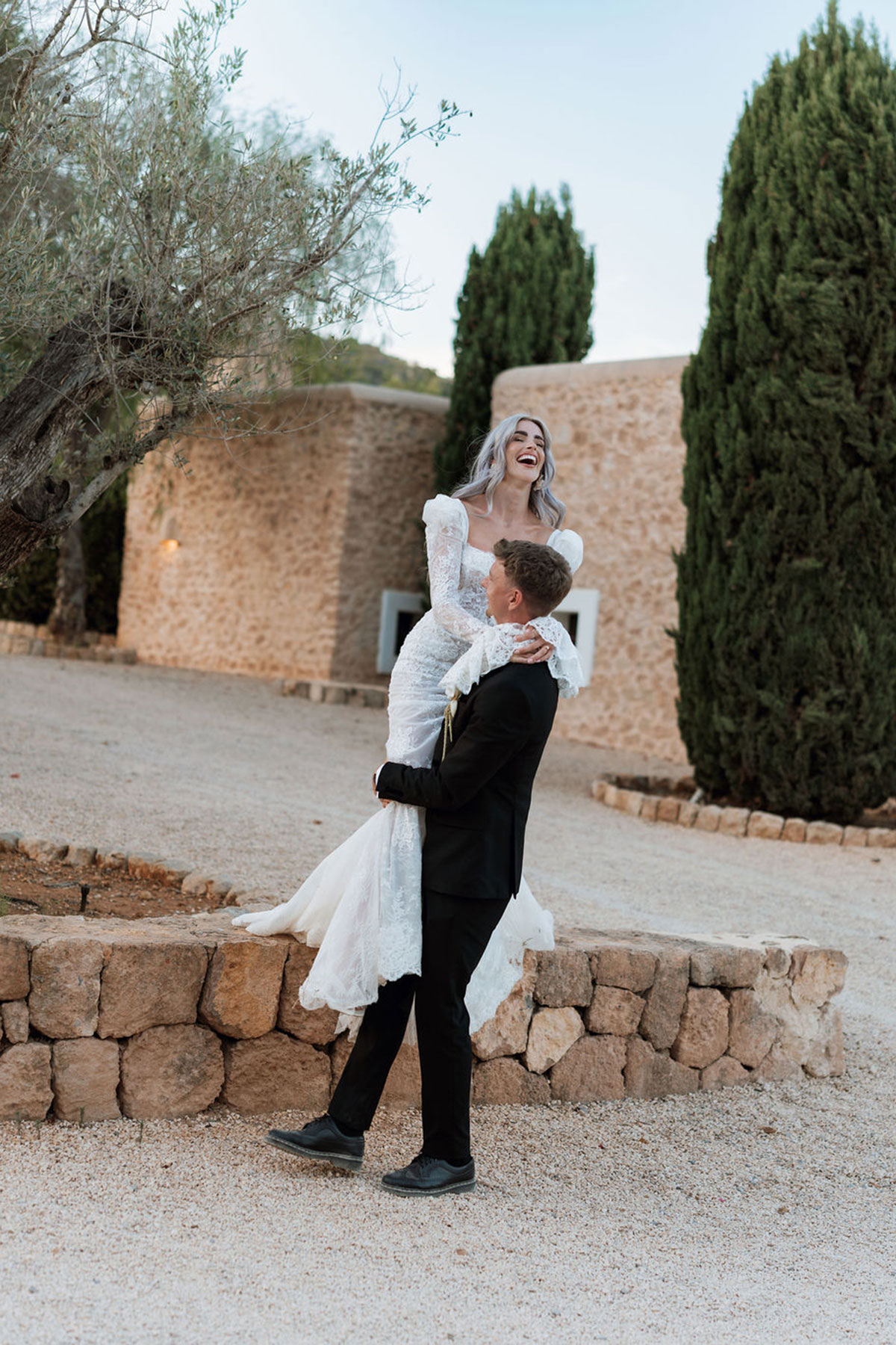 Bride in Carolyn Baxter Bridal lace wedding dress laughing as groom lifts her during sunset portraits at Can Rafal Ibiza