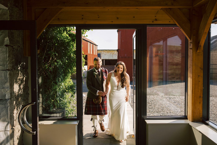A bride and groom leave a courtyard and walk through a glass door together holding hands 