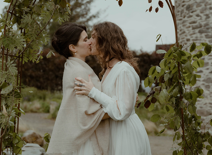 a bride wearing a shawl kissing another bride wearing a long-sleeved wedding dress. They are standing under a foliage-covered arch in a garden