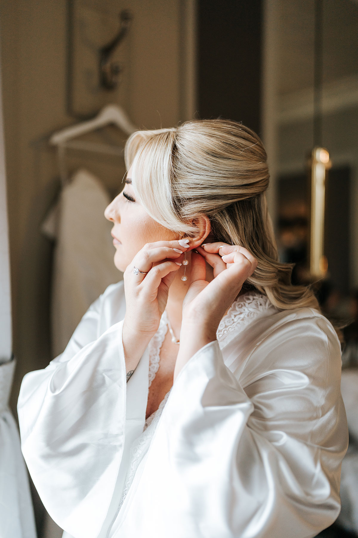 Bride in a satin robe fastening pearl drop earrings during morning preparations before her wedding ceremony
