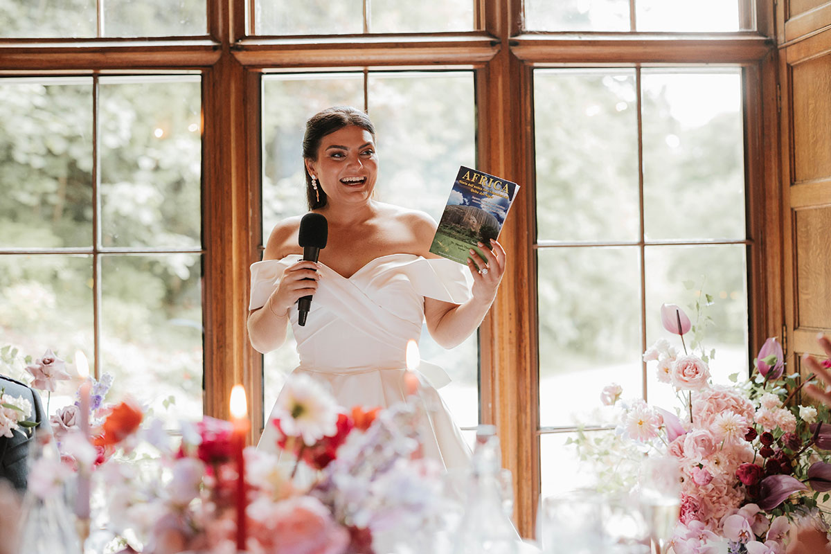 Bride giving wedding speech at Drumtochty Castle Aberdeenshire with colourful floral table décor and window backdrop