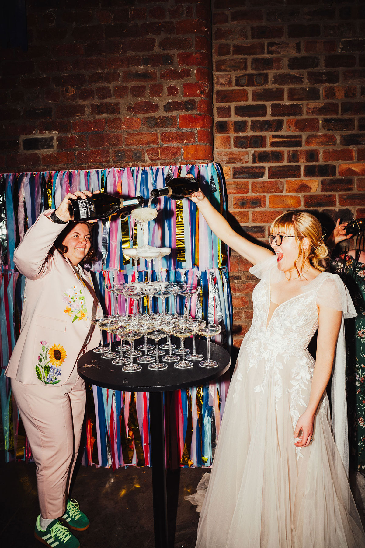 Two brides at their Engine Works wedding pouring prosecco into a stacked champagne tower, standing in front of a colourful streamer backdrop.