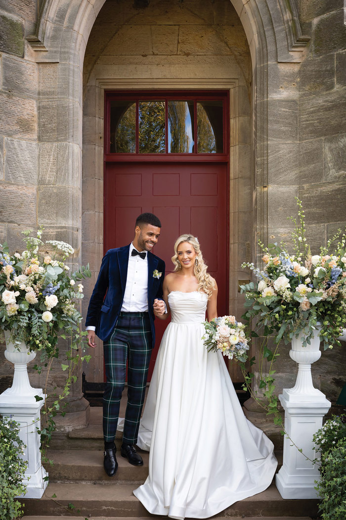 A groom wearing green tartan trousers and a navy velvet suit jacket holds hands with a bride wearing a strapless wedding dress as they walk down a set of stone steps