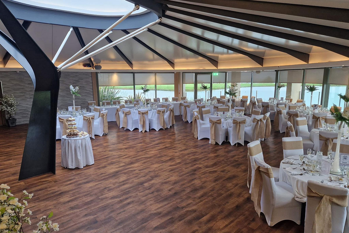 room with wooden floor has several white tablecloth set round tables with white chair covers with golden brown bows tied around the backs of all chairs and views out to the water from the room