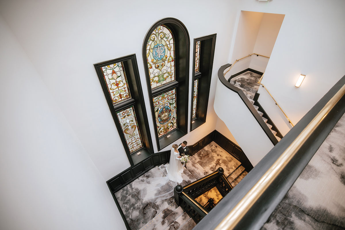 Bride and groom standing on the grand staircase beneath stained glass windows at The Collector’s Hall wedding venue in Glasgow city centre.
