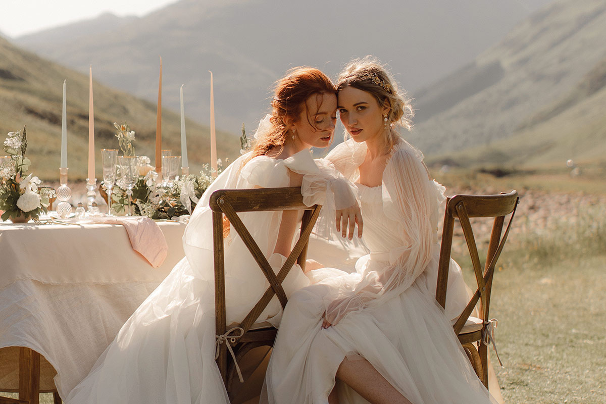 two brides sit on wooden dining chairs, leaning into each other, their tulle wedding gowns gathered around them, the tall candlesticks and glassware visible on the table and sweeping green mountains in the distance behind them