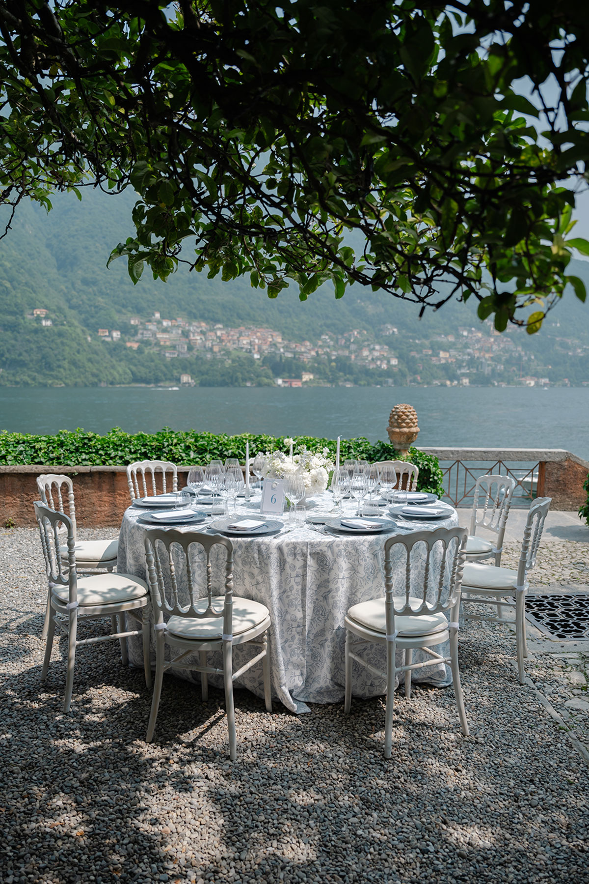Lake Como wedding table set beneath trees at Villa Regina Teodolinda, with white chairs, patterned linens and lake views in the background