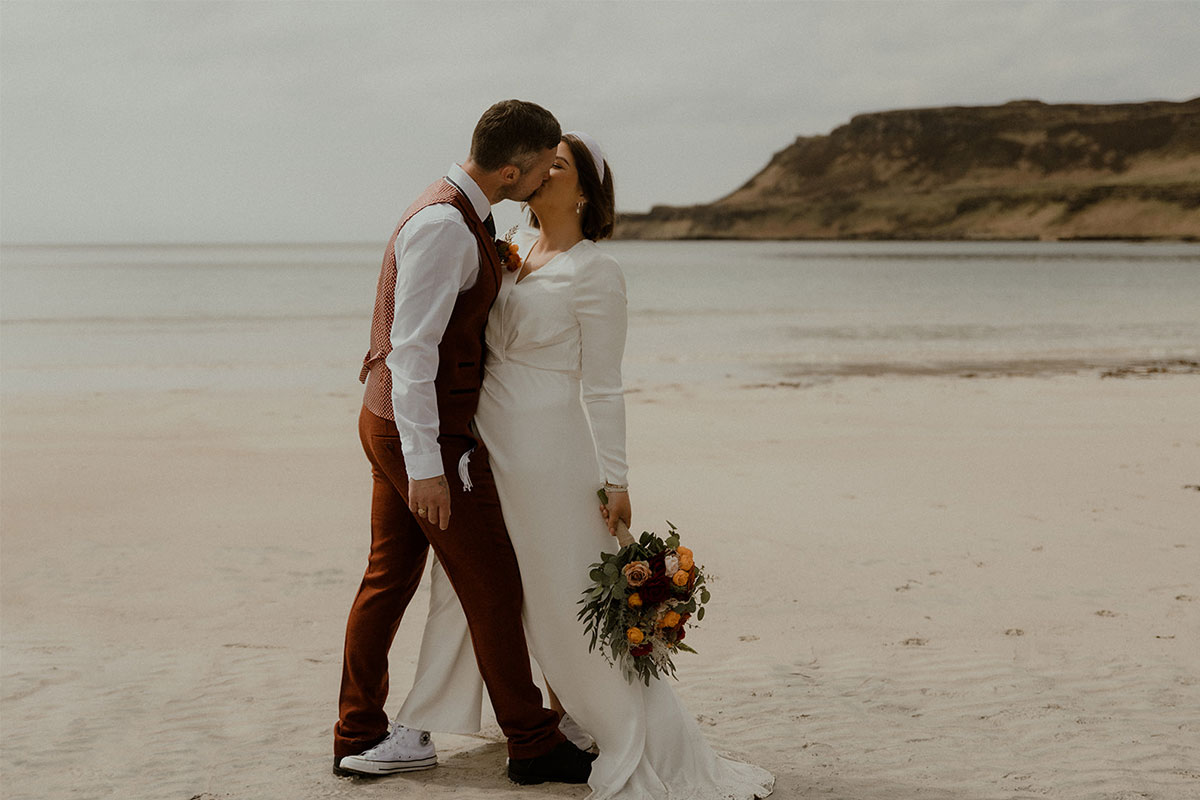 A bride and groom kiss while standing on a beach