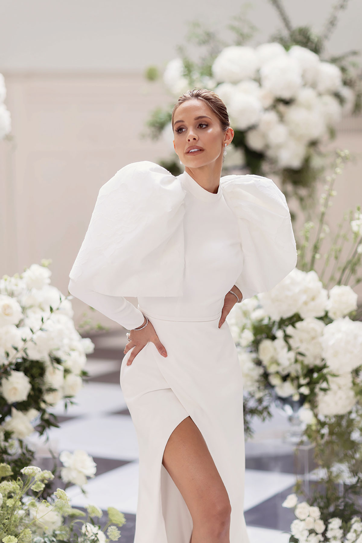 Bride in a high-neck gown with dramatic puff sleeves and thigh-split skirt, posing among white floral wedding arrangements