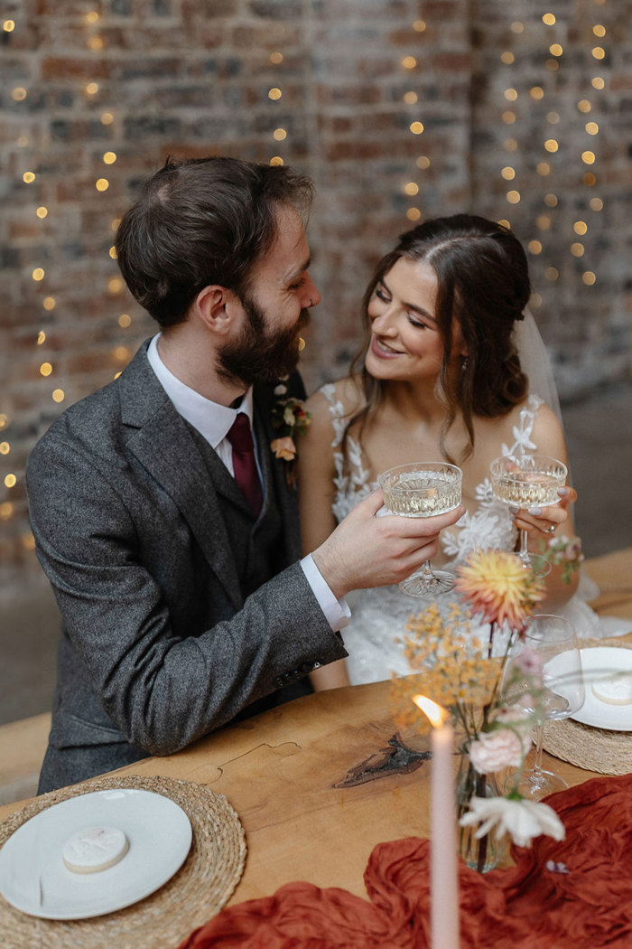 bride and groom sit beside each other on wooden dining bench, cheersing with their coupe champagne glasses while fairly lights decorate the brick wall behind them