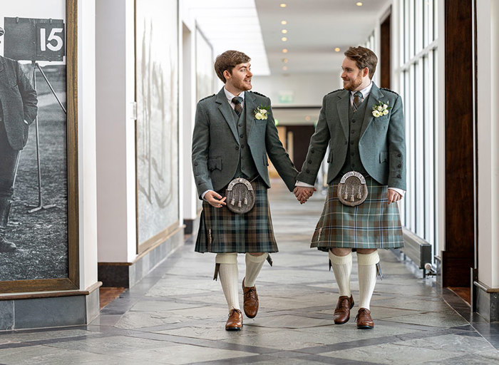 two groom wearing green tweed kilt outfits walking hand in hand along a corridor at the Old Course Hotel