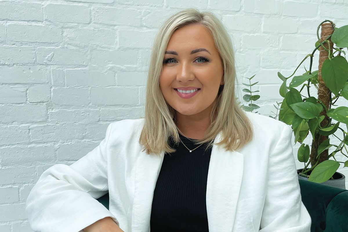 tan woman with shoulder-length blonde hair wears a white blazer and sits in front of white brick wall