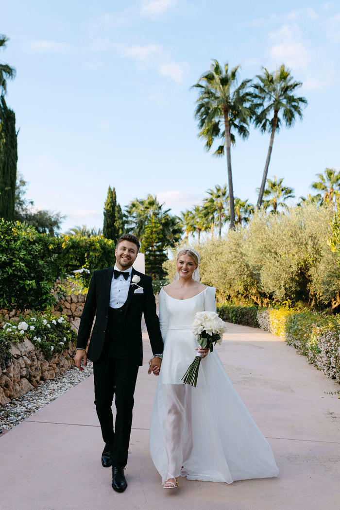 bride in long sleeve mini wedding dress with sheer overskirt and groom in tux hold hands and smile as they walk along a palm tree path