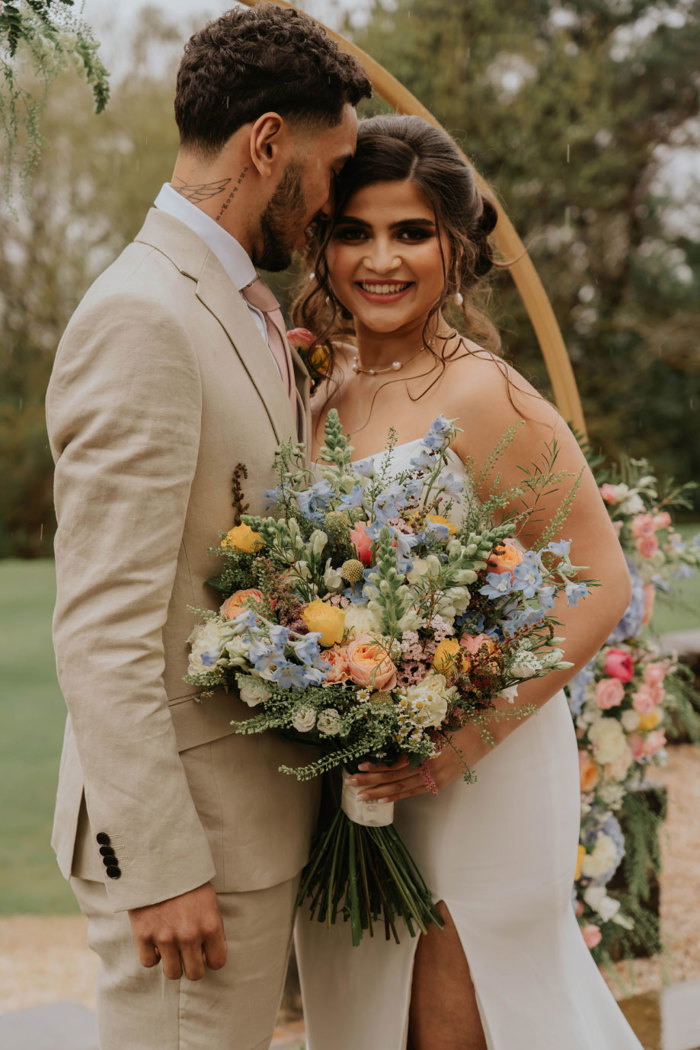 A bride holding a colourful bouquet smiles at the camera while the groom leans his head against hers