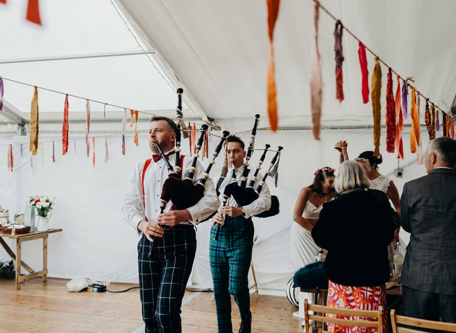 two men playing the bagpipes in a marquee that has colourful paper streamers hanging from a string either side of them