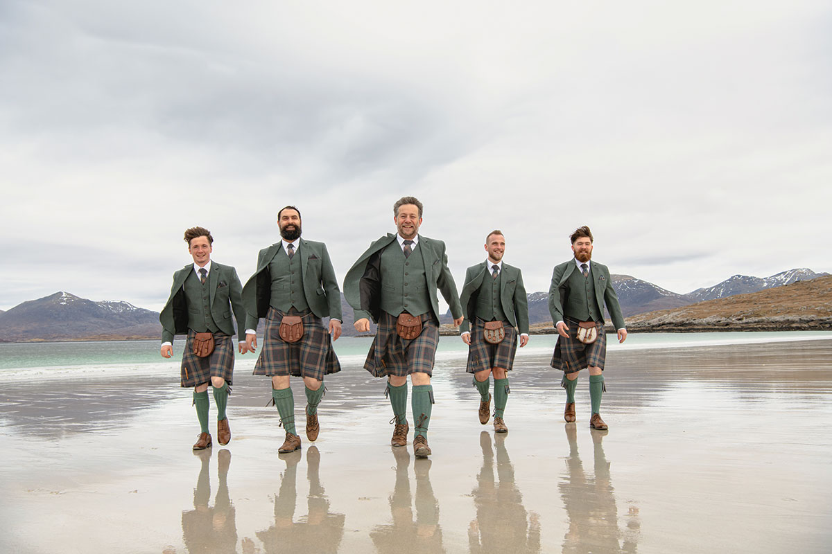 Groomsmen wearing tartan kilts and green jackets walking along a Scottish beach on a wedding day.