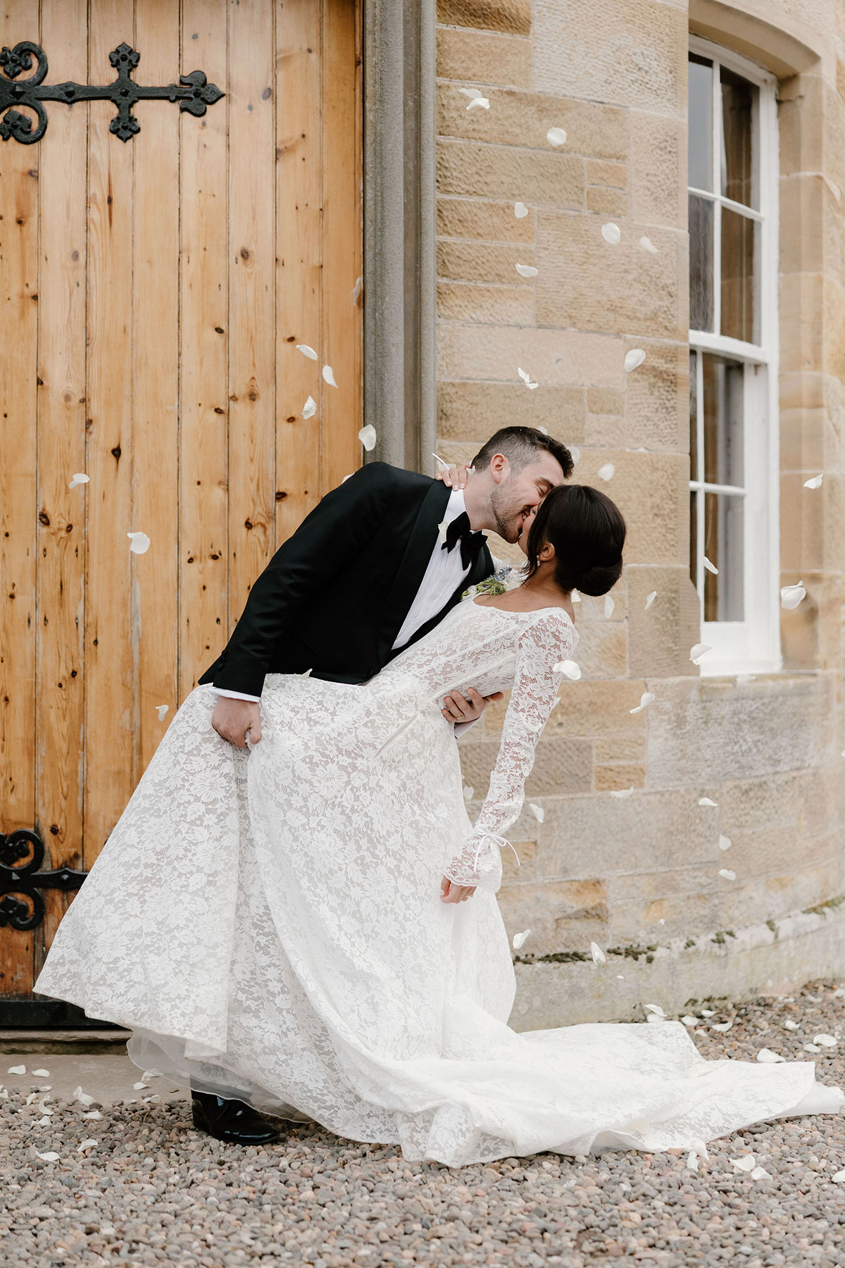 Bride and groom sharing a kiss outside a stone building as white petals fall around them