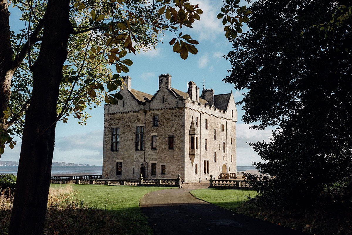 Exterior view of Barnbougle Castle near Edinburgh, framed by mature trees and gardens with coastal views in the background