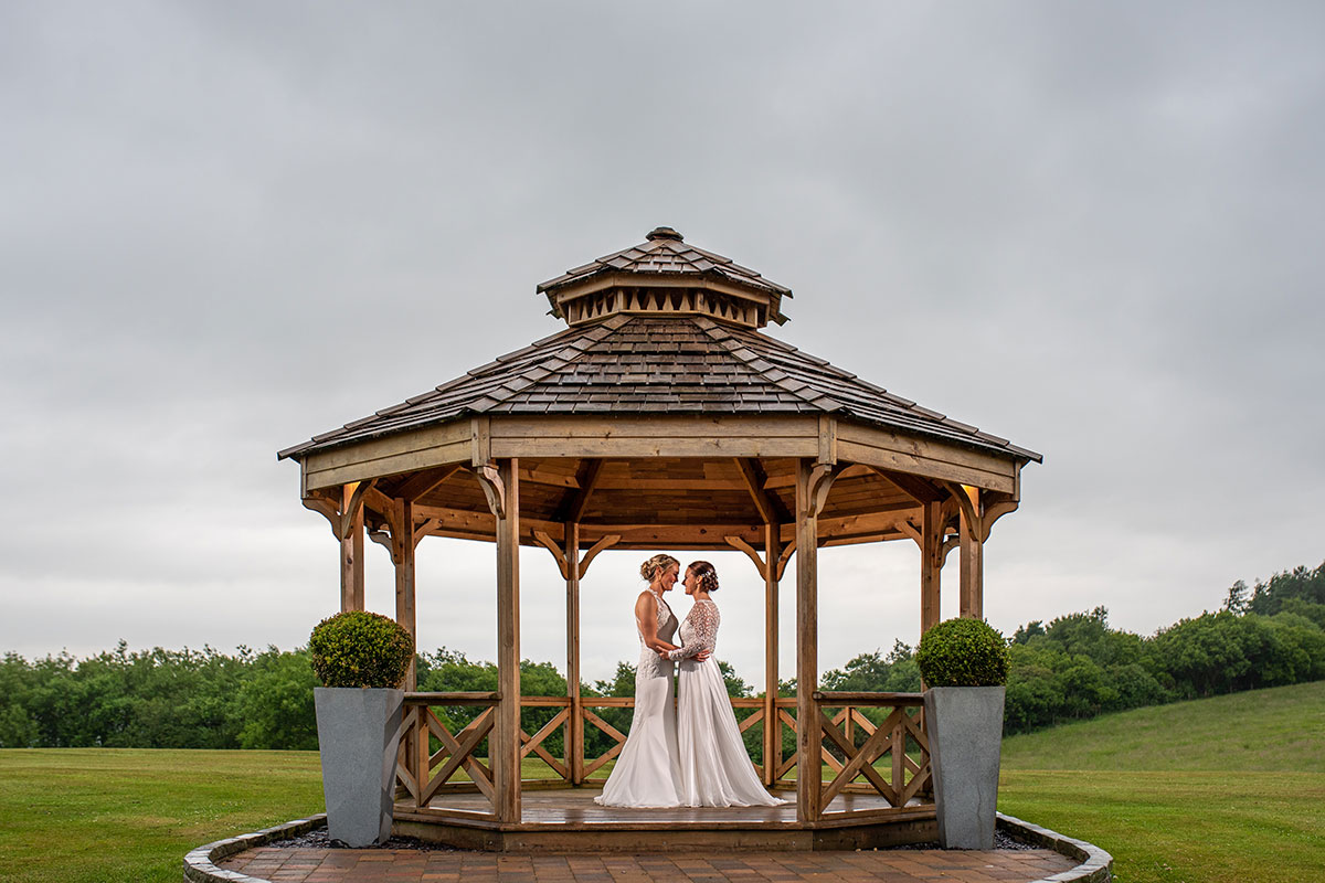 Two brides standing together beneath the wooden bandstand at The Vu wedding venue in West Lothian