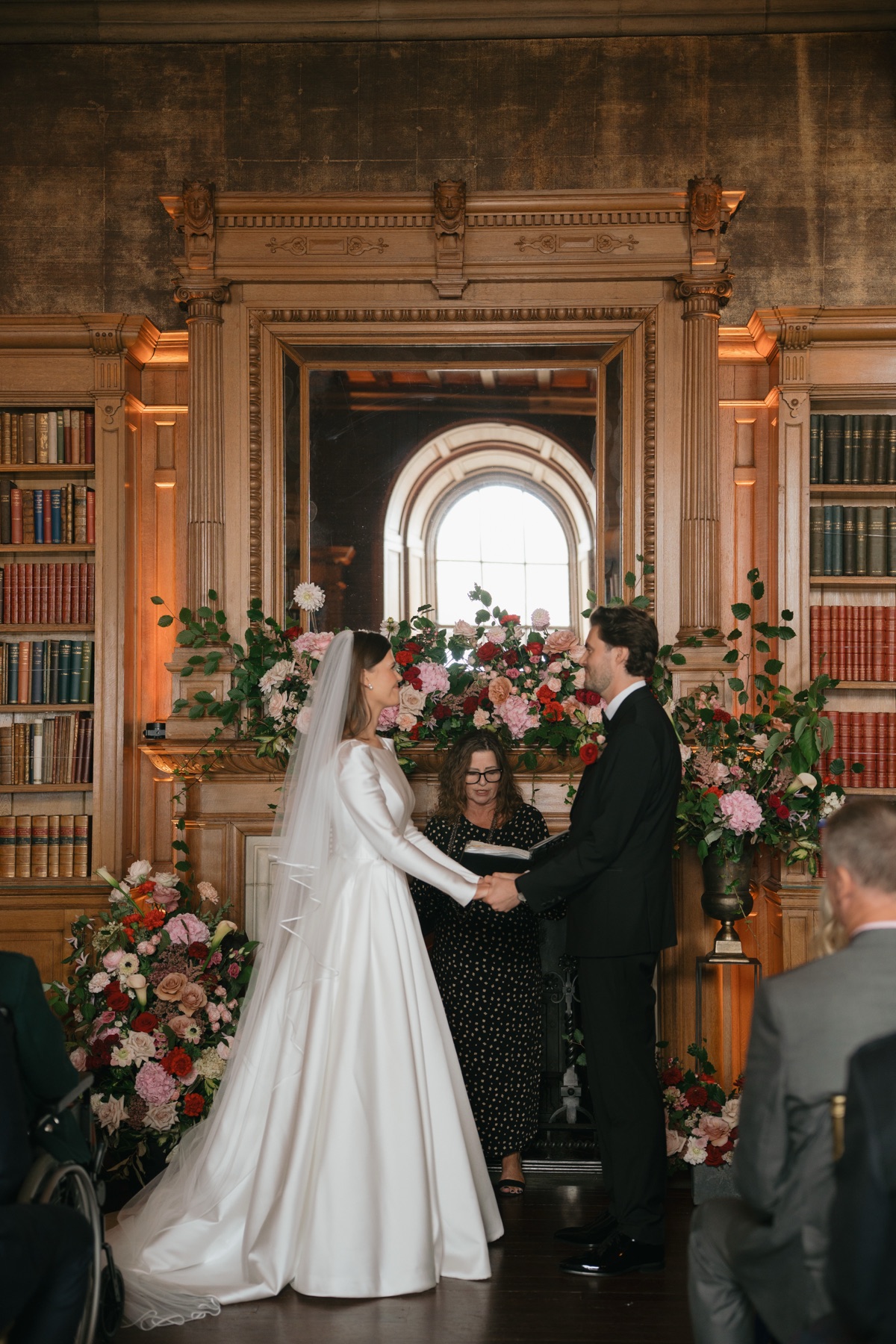 Nicole and Ryan hold hands during their wedding ceremony in the library at Barnbougle Castle, surrounded by colourful bookshelves.