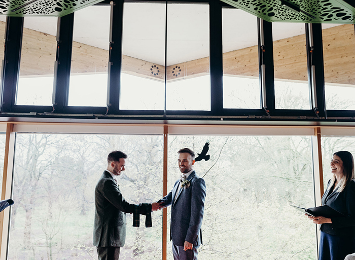 two grooms hold hands during a handfasting during their wedding ceremony in front of a large window. A smiling celebrant stands to the side and there is lots of greenery outside the window