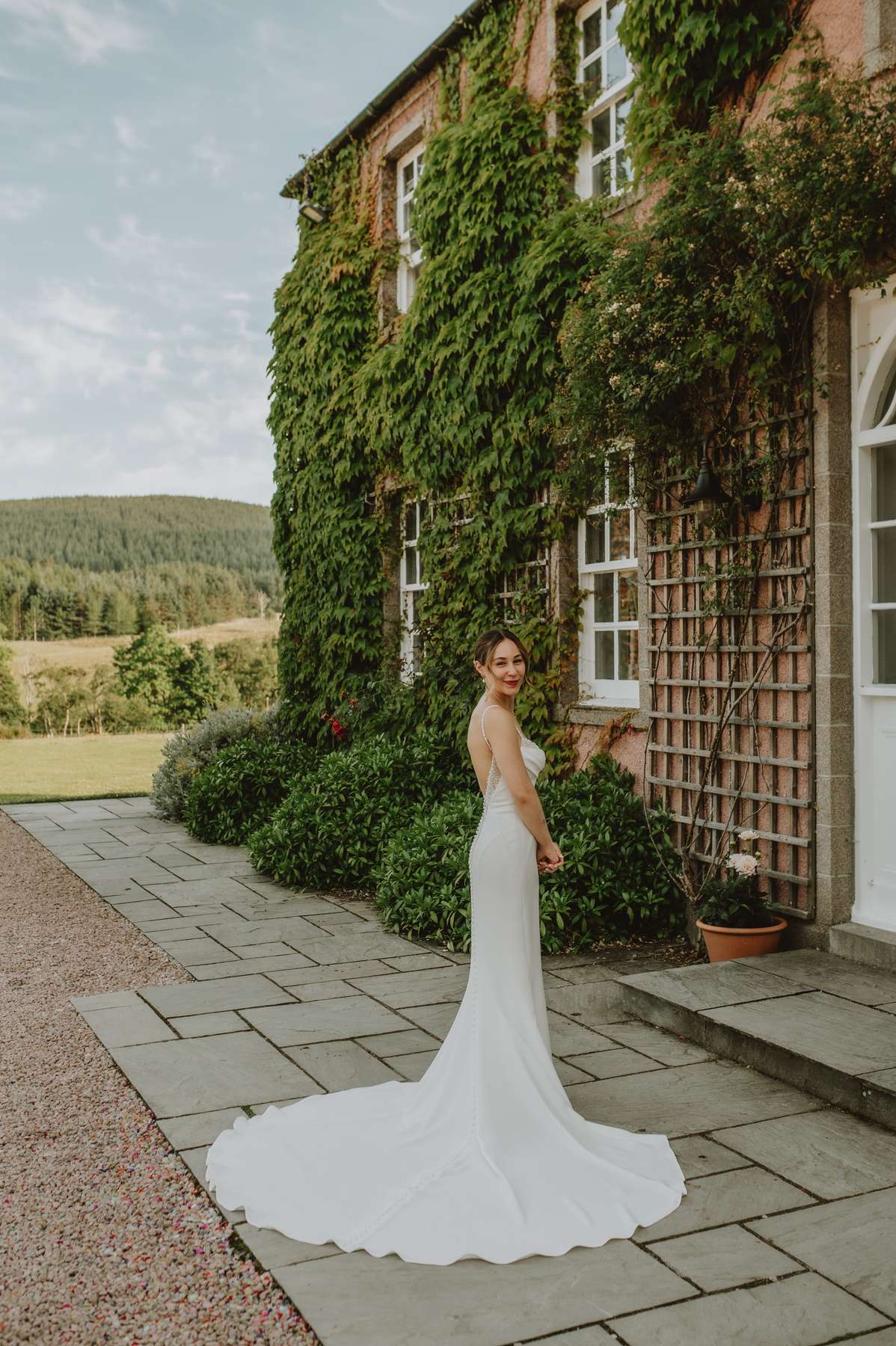 Bride in a fitted white wedding dress posing beside the ivy-covered façade of Ballogie House