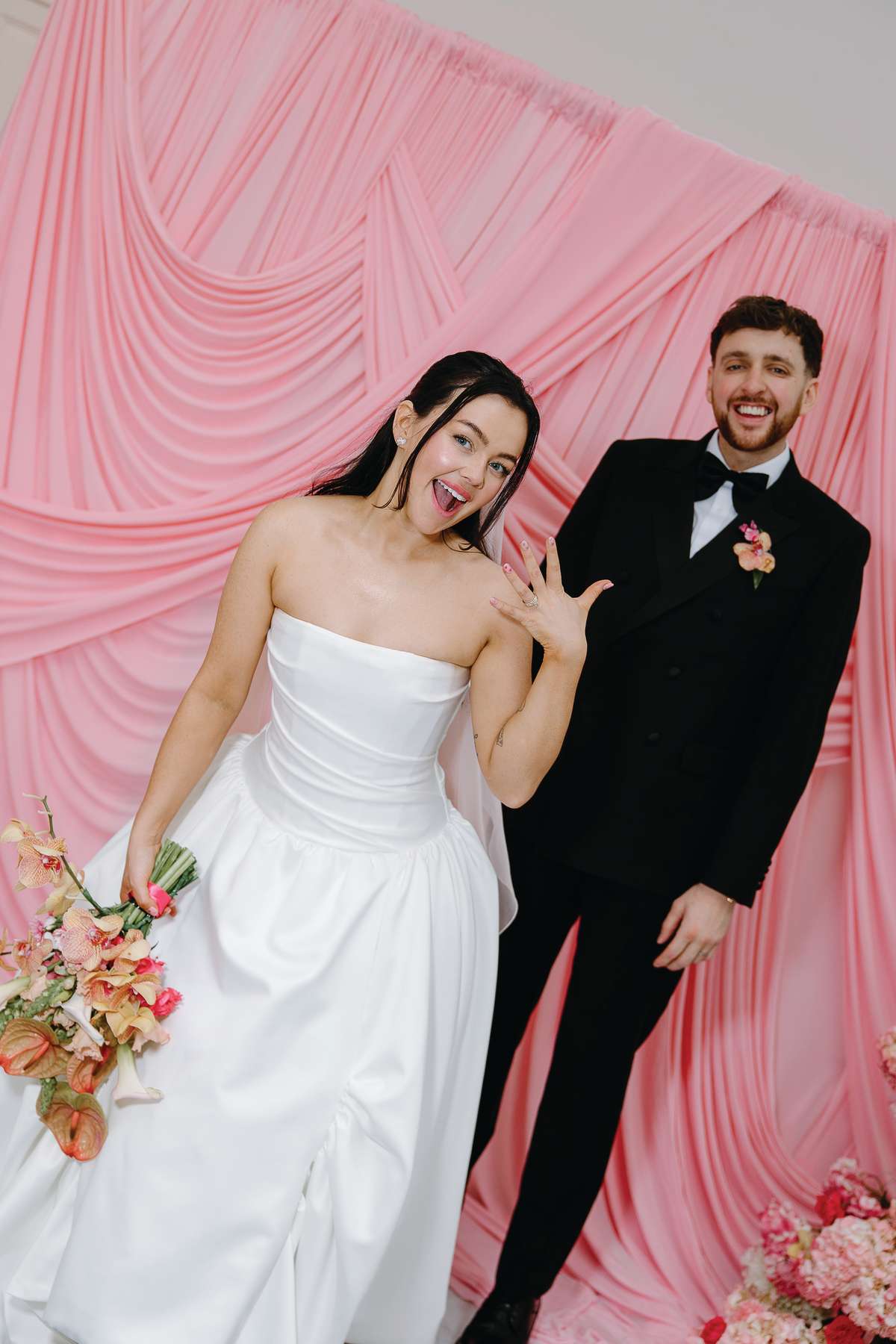 Bride shows off her wedding ring alongside the groom in front of the statement pink draped backdrop at The Collector’s Hall Glasgow.