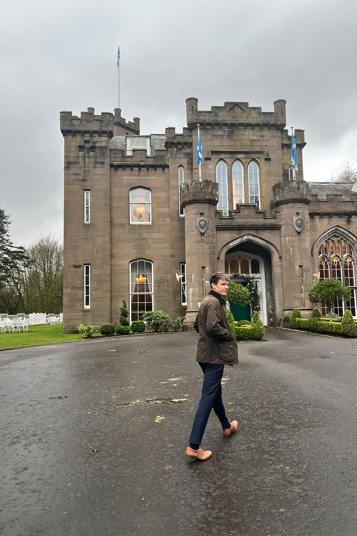 A man walking towards the entrance of a historic Scottish castle with flags flying above the turrets at dusk