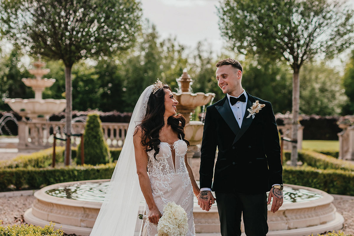 bride in v-neck wedding dress and tiara veil holds hands with groom in black tux as they both stand in front of a garden fountain