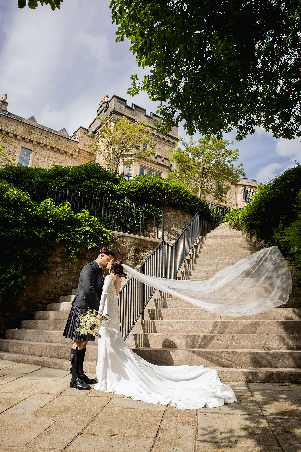 Bride and groom share a kiss on a grand stone staircase outside a castle, her long veil blowing in the breeze