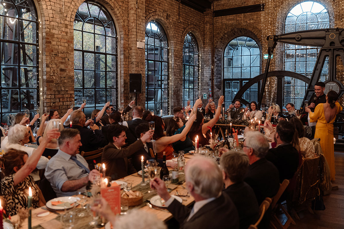 Wedding guests raise a toast during speeches in a candlelit industrial wedding reception space