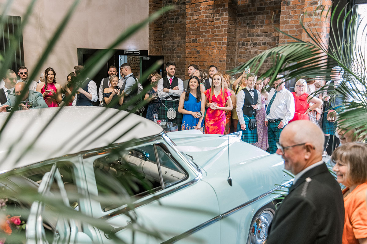 Wedding guests gather around a pale blue vintage car brought inside a brick building
