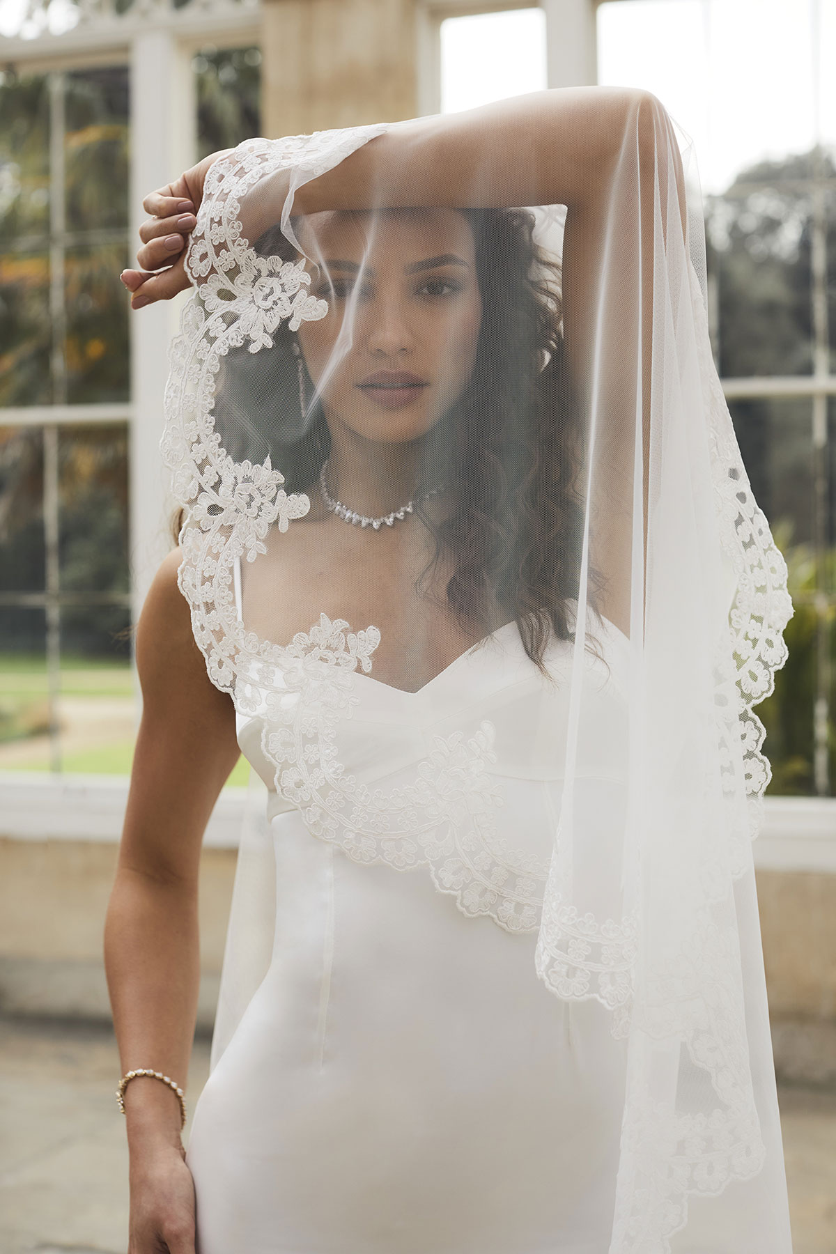 brunette curly haired woman stands in front of large windows wearing a thin strapped satin white dress and holds her one arm up above her forehead to hold up lace edged veil in front of her face