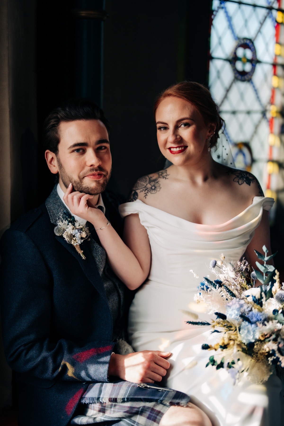Newlyweds Robyn and James pose for a wedding portrait beside a stained-glass window at Òran Mór, with Robyn holding her blue-and-white bouquet.