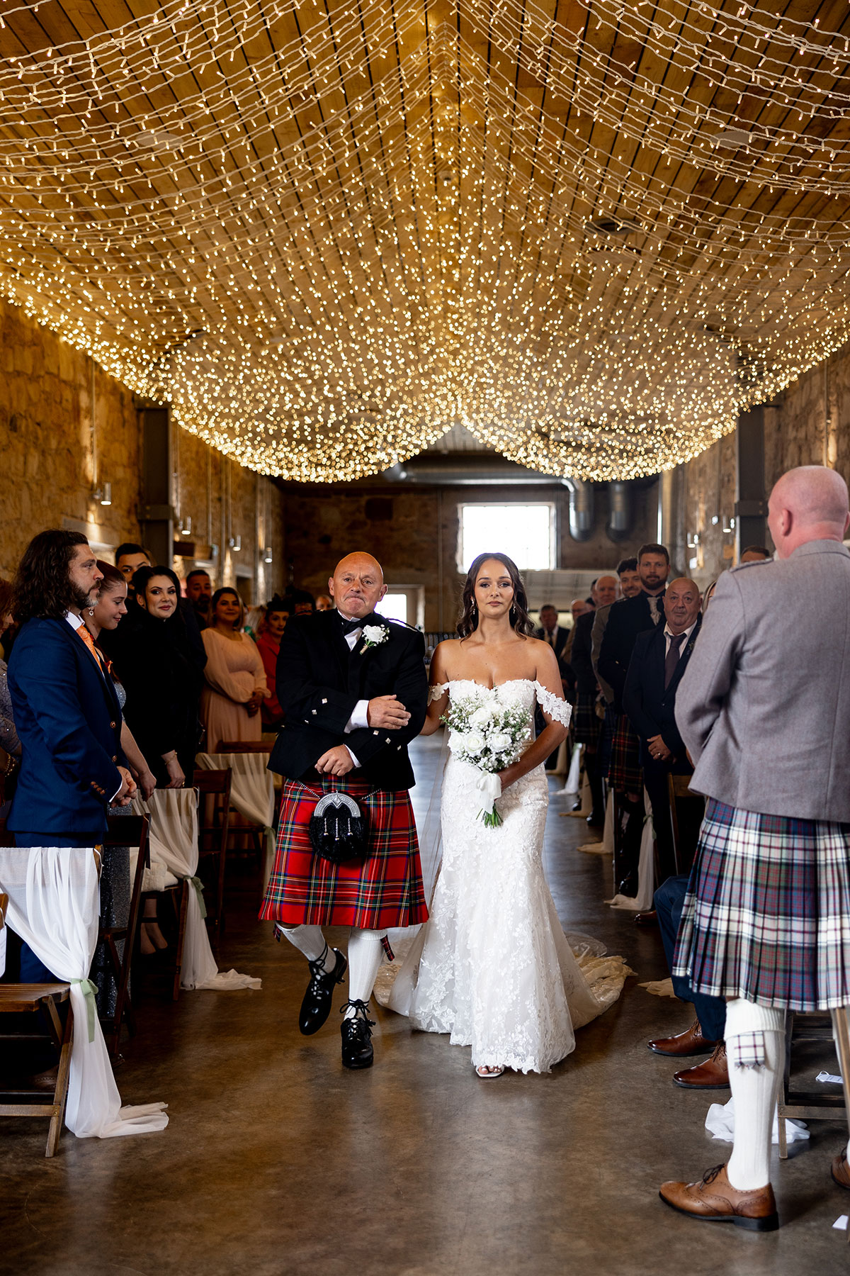 Bride walks down the aisle with her father under a canopy of fairy lights at Falside Mill barn wedding in Fife
