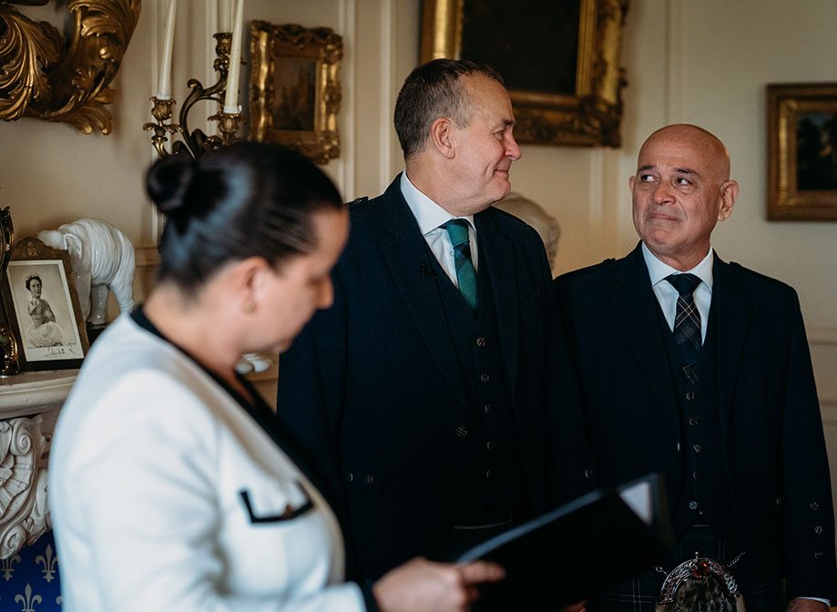 two grooms look at one another emotionally during a wedding ceremony as a person reads in front of them