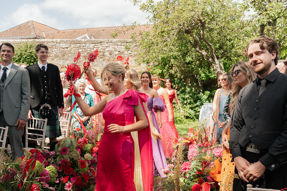 Bridesmaids dance down the flower-lined aisle carrying fans during a vibrant outdoor wedding ceremony at Kinkell Byre in St Andrews.