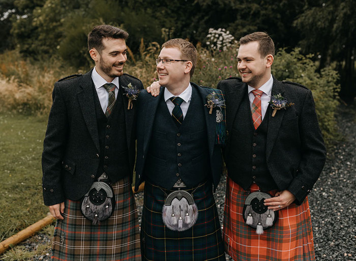 Three men in different coloured kilts look at each other as they face the camera with their arms around each other