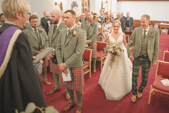 A bride walks arm in arm with her father up the aisle towards the groomsmen and officiant in a church