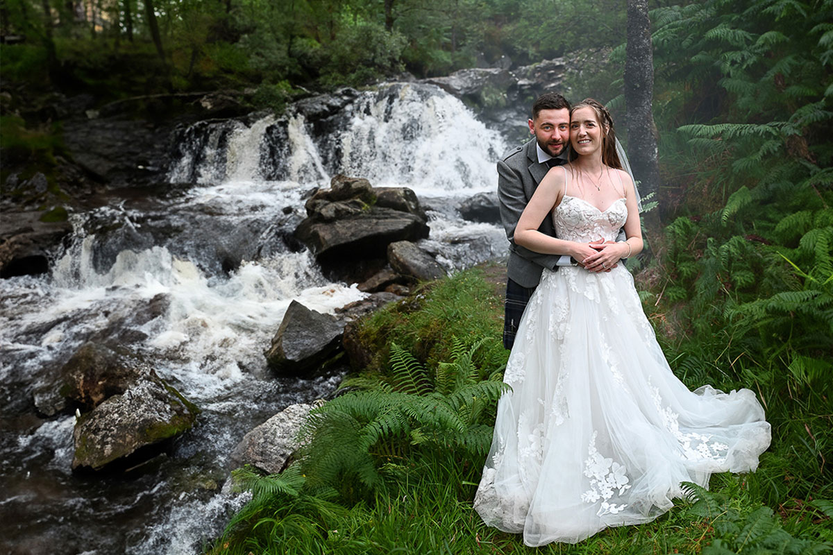 A bride and groom stand together next to a waterfall, both smiling at the camera