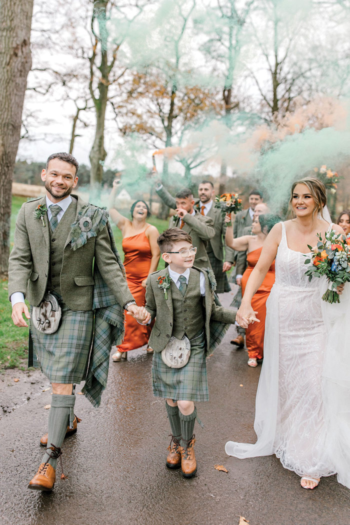 groom in blue and green tartan kilt outfit, young boy in matching kilt outfit, and bride in in beaded gown holding colourful bouquet all hold hands and walk outdoors together as colourful powder floats in the air behind them