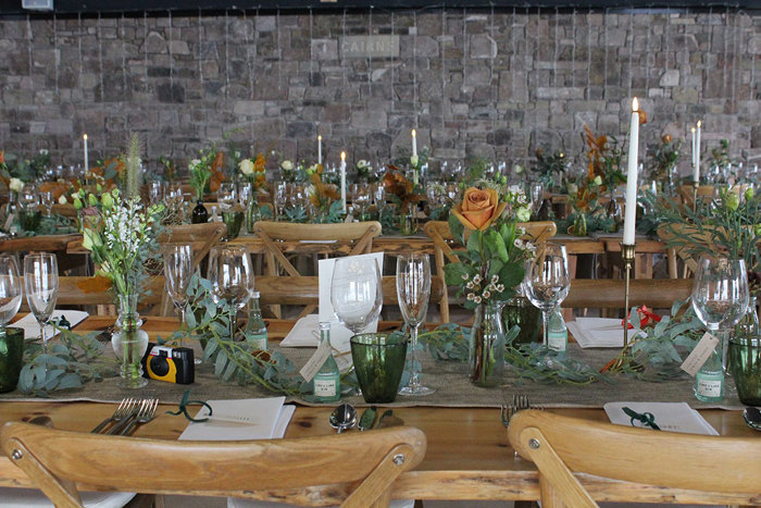 two long wooden dining tables decorated with an abundance of greenery and flowers in little vases