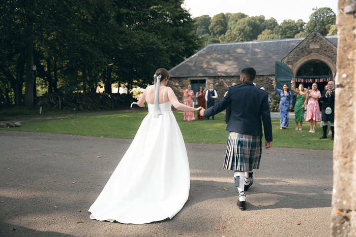 A person in a kilt and person in a wedding dress walking down a path at the Byre at Inchyra