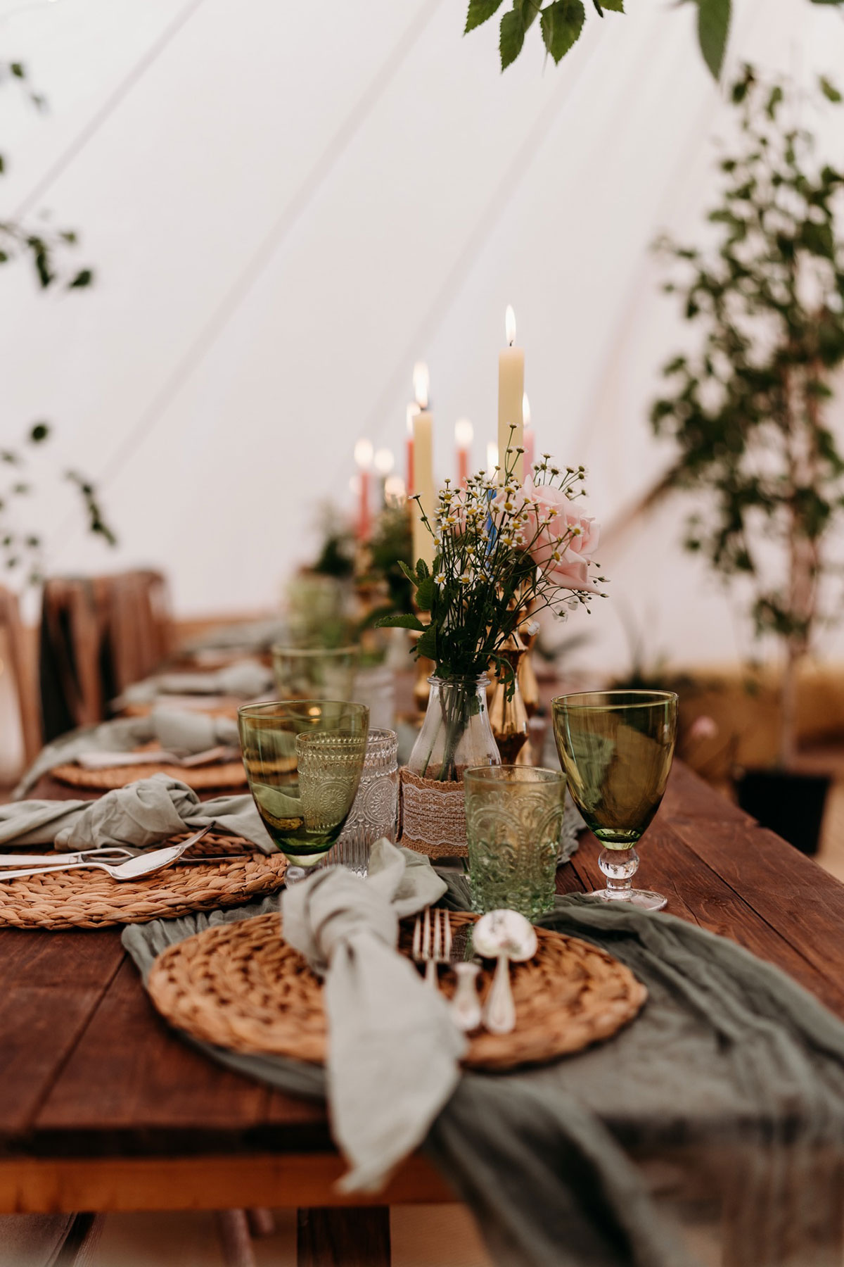 a long wooden dining table is set with rattan placemats, green napkins and glassware, bud vase flowers and colourful candlesticks