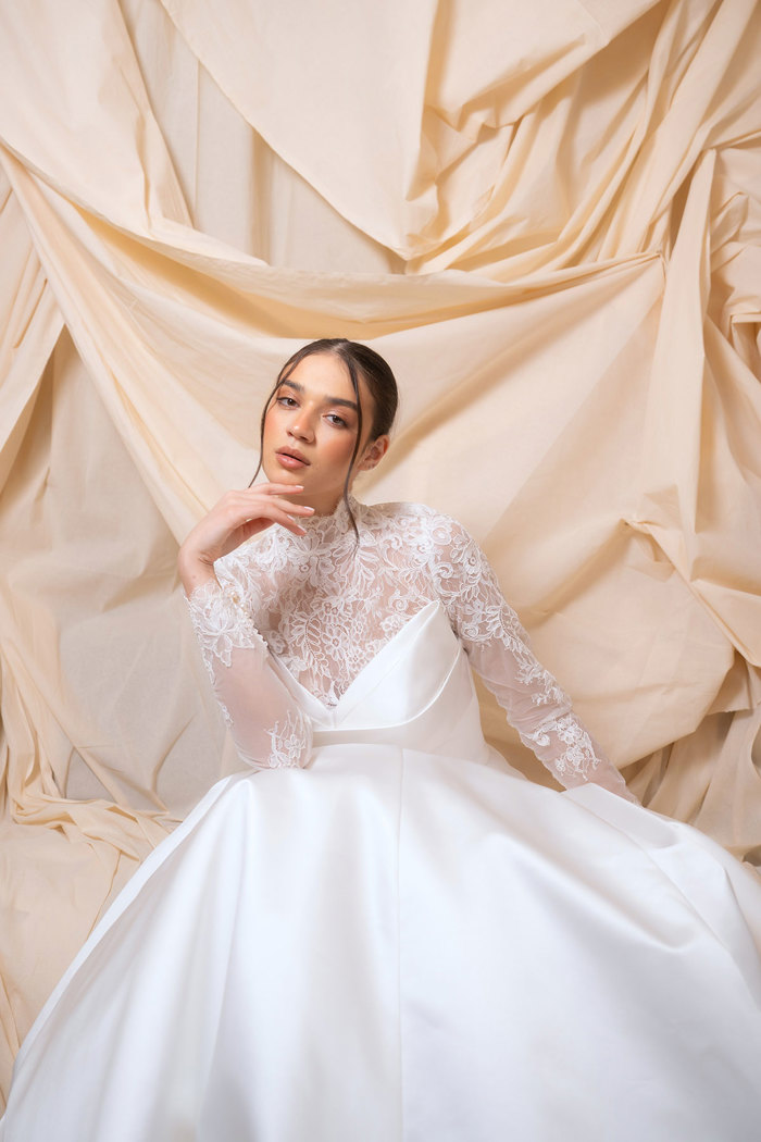 bride with dark brown hair wears lace high-neck, long sleeve top underneath strapless ballgown wedding dress, sat in front of a dark cream sheet backdrop