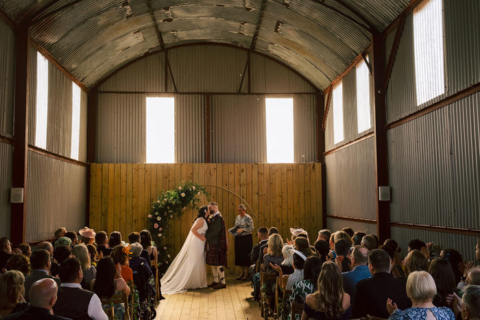 A wedding ceremony inside a barn, with the newlyweds kissing at the alter and the guests looking on