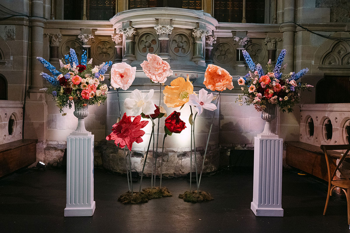 Ceremony backdrop featuring oversized paper flowers in red, peach and cream tones framed by large colourful floral arrangements on white pedestals
