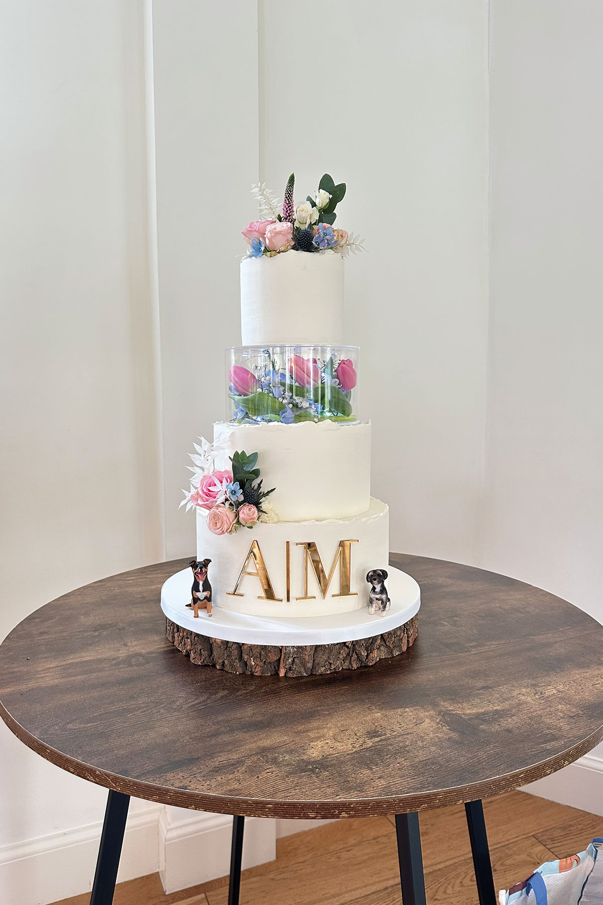 Contemporary three-tier wedding cake with fresh flowers, clear acrylic tier showcasing tulips, and gold initials “AIM”, by Jappacakes in Lanark